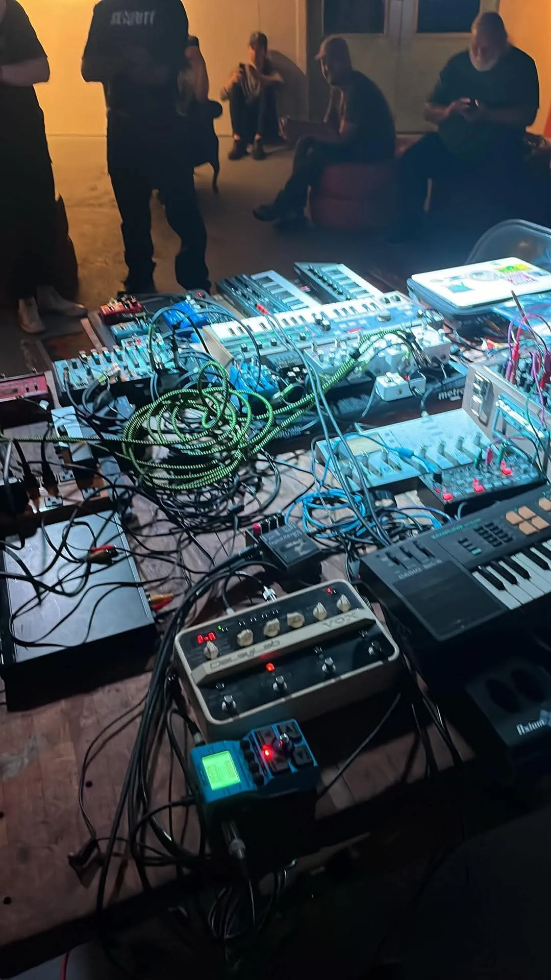 A table of synthesizers, drum machines and effects pedals tangled in green and blue patch cables, people talking in the background.